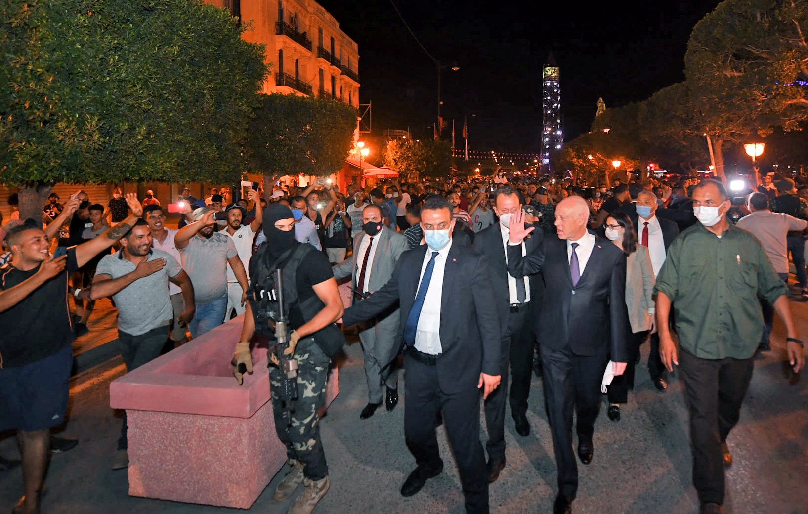 L'image montre une scène nocturne où un groupe de personnes marche dans une rue animée, entouré d'arbres et de lanternes. Au centre, on peut voir des individus en costume, accompagnés par des membres des forces de sécurité, dont l'un est vêtu de vêtements sombres et porte un masque. La foule semble exprimée une certaine agitation, avec des gestes de la main et des expressions variées sur les visages. L'atmosphère générale est tense, et il y a un contraste entre les personnes en tenue formelle et le cadre plus informel de la mobilisation populaire.