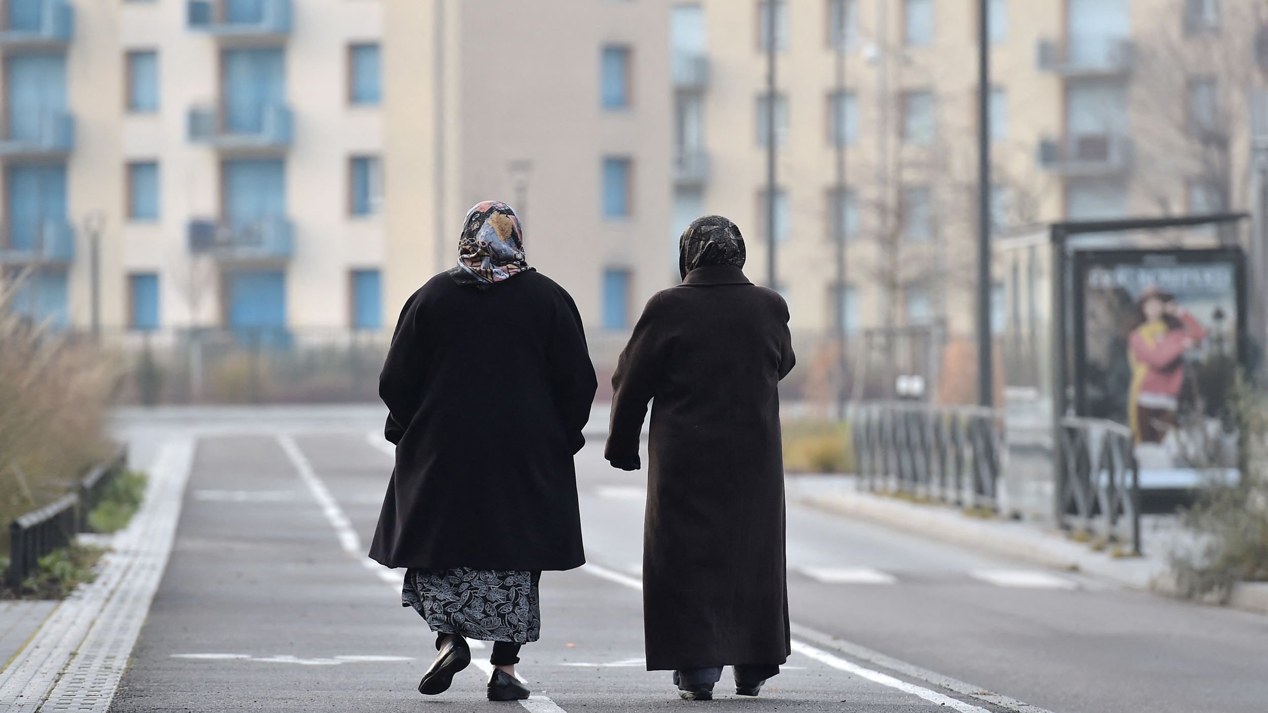 L'image montre deux femmes marchant côte à côte sur une voie urbaine. Elles portent des manteaux sombres et des foulards, ce qui suggère qu'il fait frais. En arrière-plan, on distingue des bâtiments modernes avec des balcons et des fenêtres. La scène dégage une atmosphère tranquille, avec un chemin bordé d'herbes. Leurs silhouettes semblent illustrer un moment de complicité et de camaraderie.