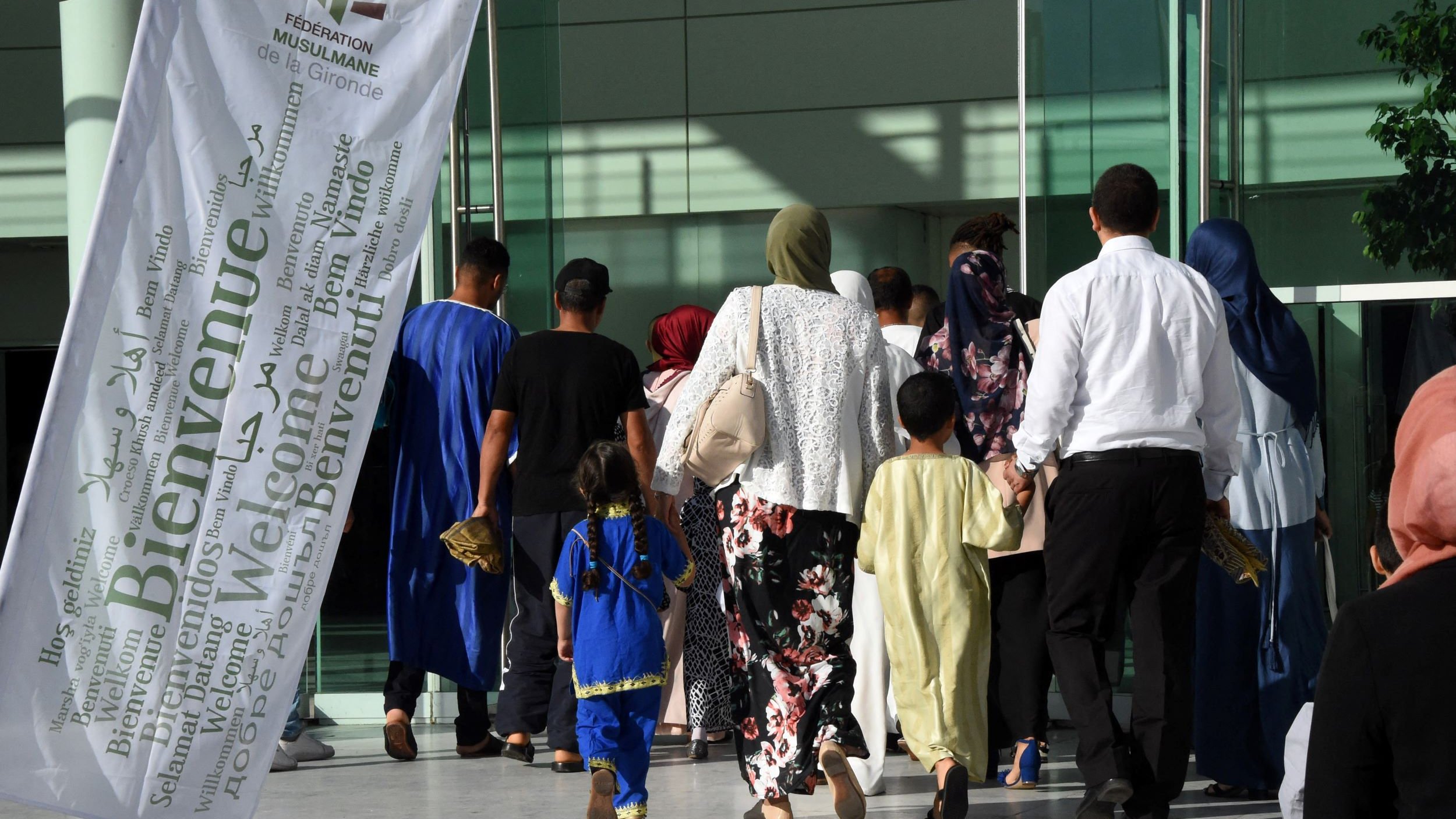L'image montre un groupe de personnes se dirigeant vers un bâtiment. On peut voir des individus de différentes générations, y compris des enfants. Certains portent des vêtements traditionnels, et plusieurs femmes ont des foulards. Il y a également une grande bannière blanche flottant au vent, portant le mot "Bienvenue" en plusieurs langues. L'ambiance semble chaleureuse et accueillante.