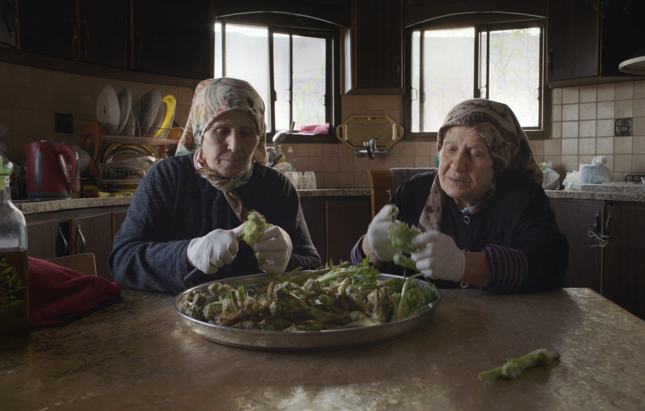 L'image montre deux femmes assises à une table dans une cuisine, en train de préparer des légumes. Elles portent des foulards et des gants blancs. Sur la table, il y a un grand plat rempli de légumes que les femmes épluchent ou nettoient. La cuisine a un décor simple avec des fenêtres qui laissent passer la lumière naturelle. L'atmosphère semble chaleureuse et conviviale, mettant en avant un moment de partage et de tradition culinaire.