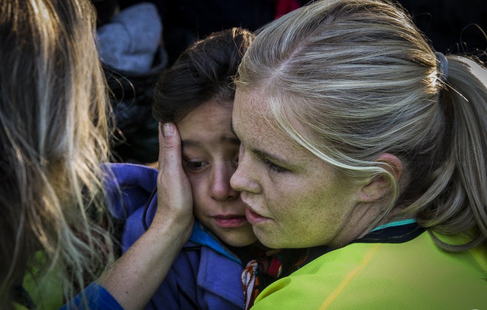 L'image montre une scène émotive où une femme, vêtue de vêtements de couleur vive, réconforte une petite fille. La femme semble douce et protectrice, tenant délicatement le visage de l'enfant. La petite fille a l'air triste ou inquiète, et elle se tourne vers la femme pour chercher du réconfort. L'environnement semble être chargé d'émotion, avec des personnes présentes en arrière-plan. Cette interaction illustre un moment de soutien et de compassion.
