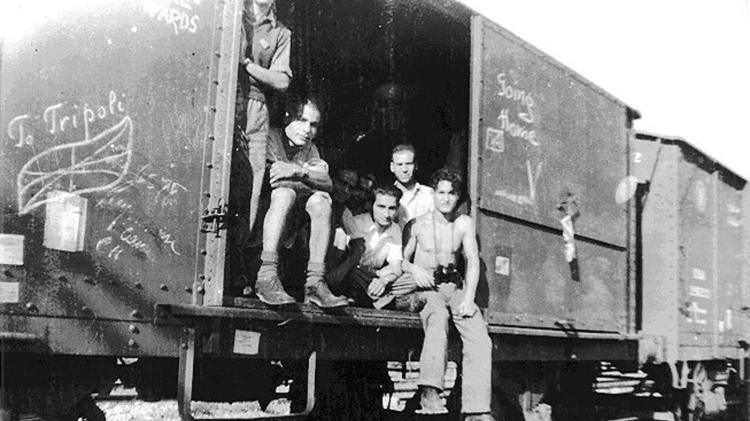 تظهر الصورة مجموعة من الأشخاص يجلسون داخل عربة قطار، حيث يبدو أنهم متجمعون معًا. الخلفية تتكون من السكك الحديدية وأحد العوارض، بينما العربة تحمل بعض الكتابات أو الرسومات على الجوانب. يعكس المشهد إحساسًا بالتاريخية والانتماء، وقد يكون له علاقة بفترة معينة في الزمن.