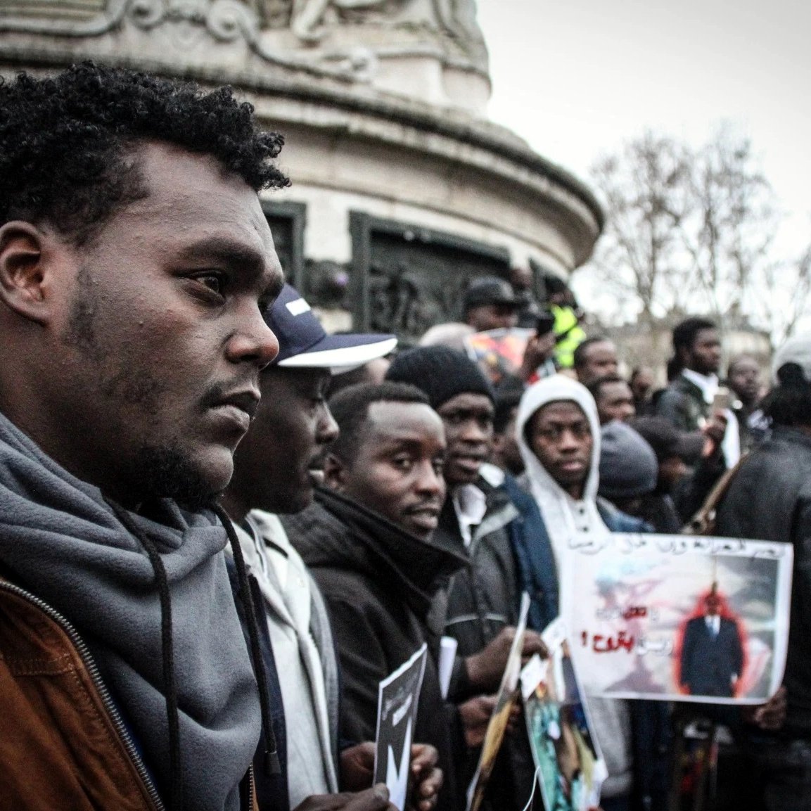 L'image montre un groupe de personnes rassemblées, probablement lors d'une manifestation ou d'un événement public. Au premier plan, un homme au port de tête pensif se distingue, entouré d'autres participants qui tiennent des pancartes et des photos. L'atmosphère semble engagée et solidaire, avec des affiches qui pourraient exprimer des revendications ou des messages de protestation. L'environnement urbain est visible en arrière-plan, suggérant que cet événement se déroule dans un espace public.