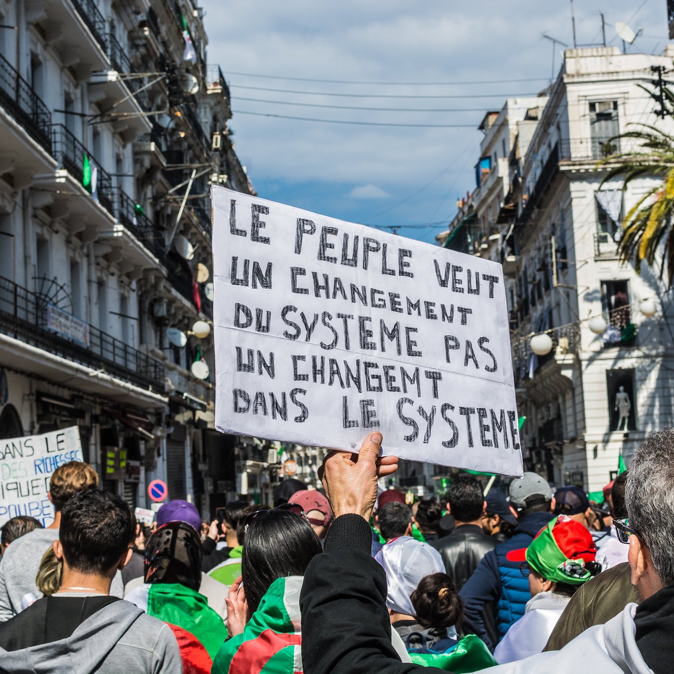 L'image montre une manifestation dans une rue animée. Au premier plan, une personne tient une pancarte qui exprime un message clair : "Le peuple veut un changement du système pas un changement dans le système". On aperçoit des manifestants qui portent des drapeaux et semblent mobilisés. Les bâtiments environnants évoquent une ambiance urbaine et dynamique, tandis que le ciel est partiellement nuageux. L'ensemble traduit une forte volonté collective de changement.