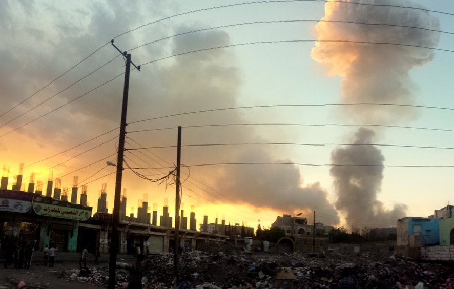 L'image montre un paysage urbain, probablement dans une zone affectée par des conflits. On peut voir des colonnes de fumée s'élevant dans le ciel, indiquant une explosion ou un incendie. À l'arrière-plan, le soleil se couche, créant un contraste entre les couleurs chaudes du ciel et les fumées sombres. Au premier plan, il y a des débris et des déchets éparpillés, et des bâtiments aux murs usés, ornés d'affiches et de signes en arabe. Les fils électriques traversent la scène, renforçant l'impression d'un environnement urbain chaotique.