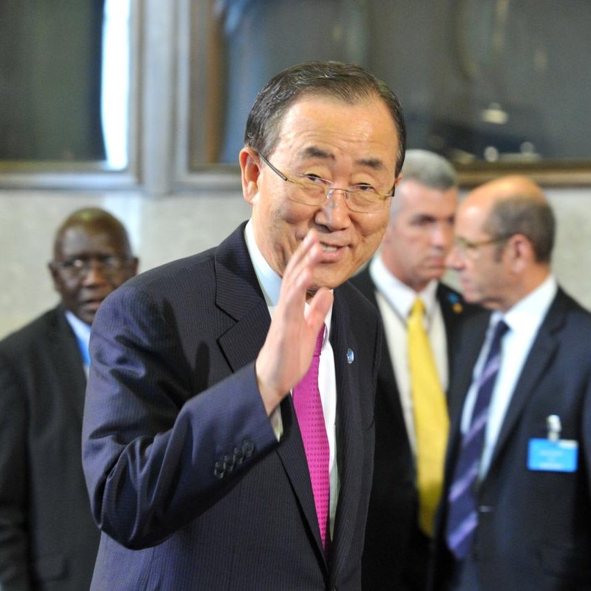 L'image montre un homme souriant, vêtu d'un costume sombre avec une cravate rose. Il semble faire un geste de la main, comme s'il saluait. En arrière-plan, on peut apercevoir d'autres hommes en costume, certains portant des badges. L'environnement semble être un lieu formel, probablement dans un contexte de réunion ou de conférence internationale.