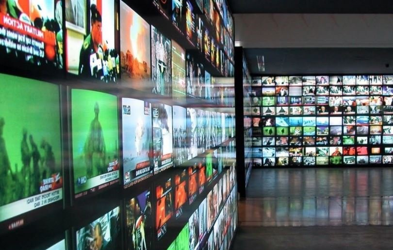 L'image montre une vaste installation composée de nombreux écrans de télévision affichant différentes vidéos et images. Les écrans sont regroupés sur des murs, créant une mosaïque visuelle. Certaines chaînes semblent diffuser des nouvelles, tandis que d'autres présentent des contenus variés. L'éclairage est vif, et l'ensemble donne une impression de dynamisme et de surcharge d'information. L'environnement pourrait évoquer un espace dédié aux médias ou à l'art numérique.