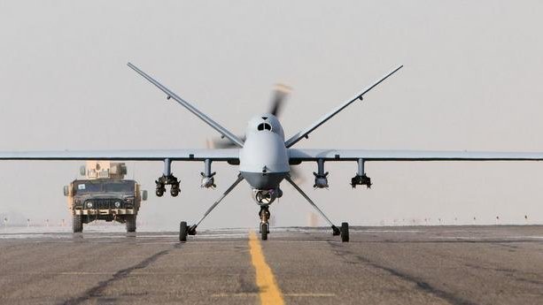 L'image montre un drone militarisé en position de décollage, vu de face, sur une piste d'atterrissage. En arrière-plan, on aperçoit un véhicule militaire qui s'approche. Le ciel semble légèrement nuageux ou brumeux, créant une ambiance de calme avant l'action. Le drone est de couleur claire, avec des ailes larges et un design aérodynamique, prêt à s'envoler.