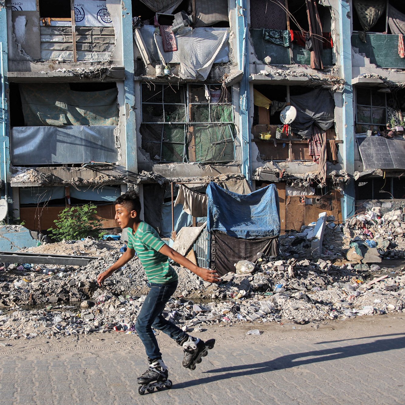 در تصویر یک پسر بچه دیده می‌شود که با اسکیت روی سطحی صاف در حال حرکت است. در پس‌زمینه، ساختمان‌هایی با نمای خراب و آسیب‌دیده قرار دارند که پنجره‌هایشان با پارچه و مواد دیگر پوشانده شده است. اطراف ساختمان مملو از زباله و خرابه است که نشان‌دهنده وضعیت دشوار منطقه است. فضای کلی تصویر حس نابه‌سامانی و فقر را منتقل می‌کند.