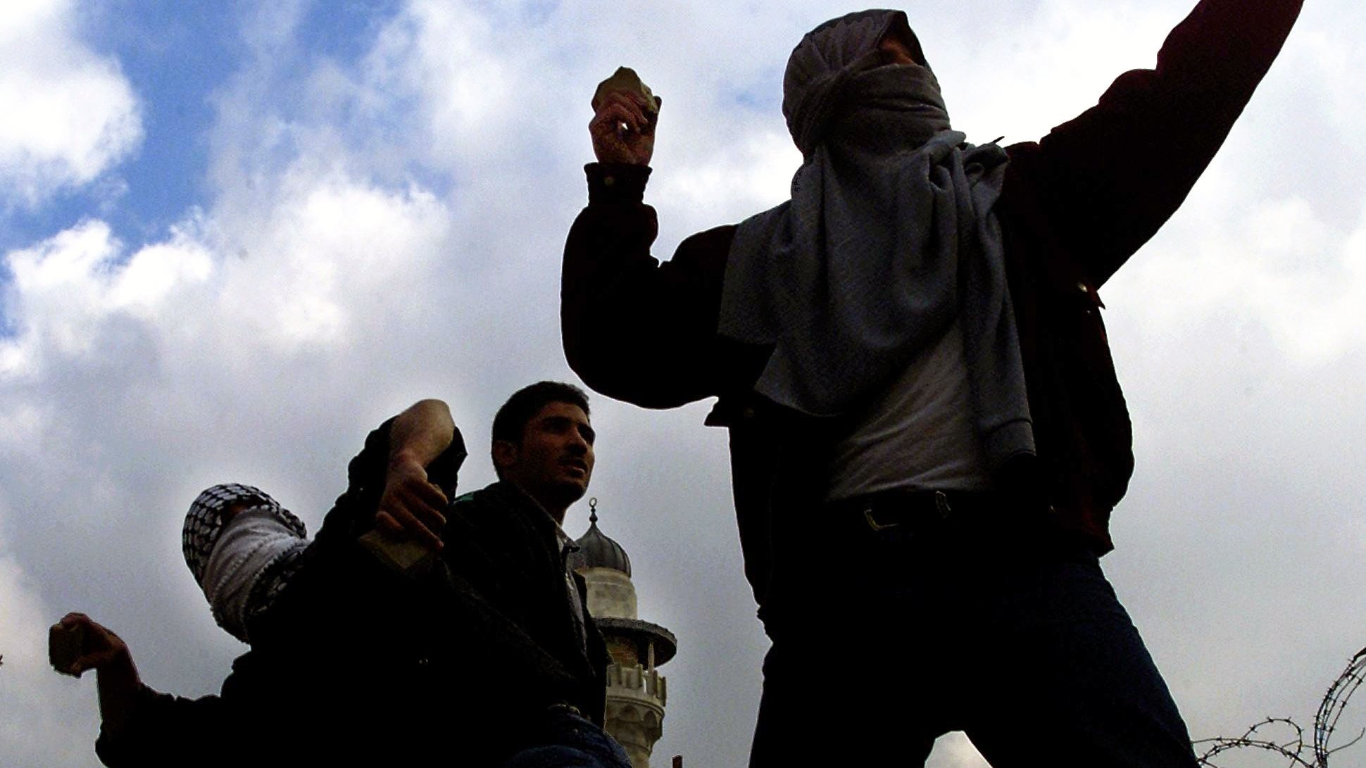 L'immagine mostra un gruppo di persone in una situazione di tensione, probabilmente in un contesto di protesta o conflitto. Alcuni individui sono coperti da sciarpe o fazzoletti, mentre sembrano lanciare dei proiettili, forse pietre. Sullo sfondo si possono vedere strutture architettoniche e un cielo nuvoloso, che contribuiscono all'atmosfera drammatica della scena. La presenza di filo spinato suggerisce una situazione di conflitto o di controllo militare.