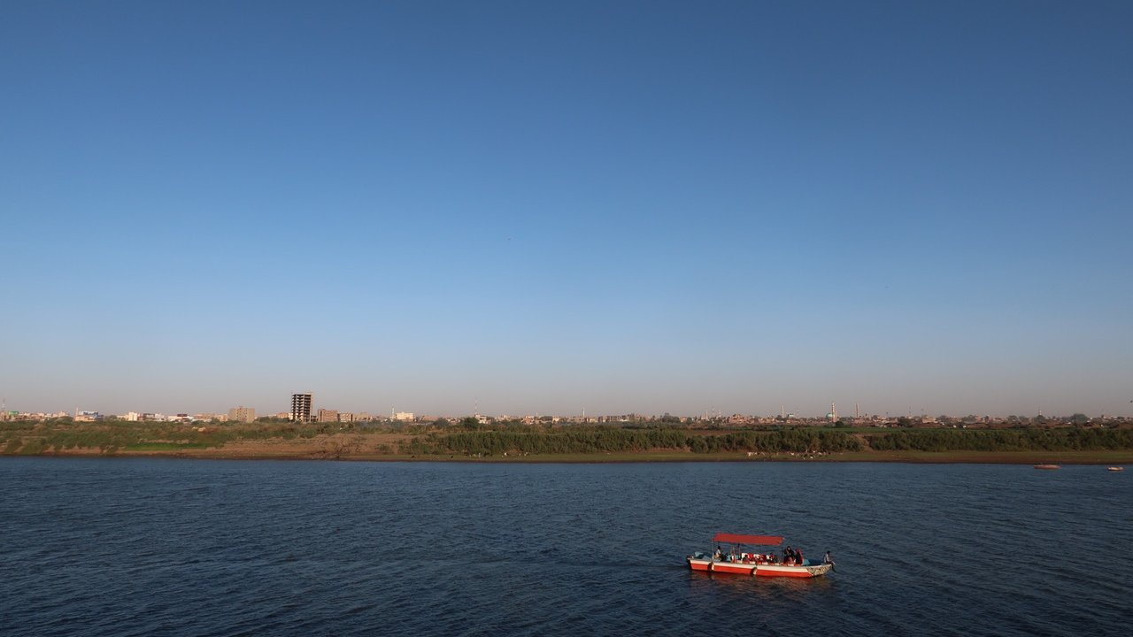 The image depicts a serene river landscape under a clear blue sky. In the foreground, a small boat with a red canopy is floating on the water, which reflects the sky. In the background, there is a sandy shore lined with greenery, and a distant cityscape can be seen, with buildings rising against the horizon. The overall scene conveys a peaceful and picturesque atmosphere.