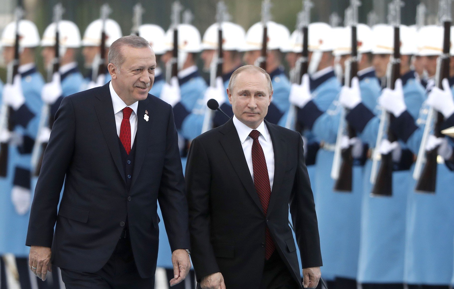 L'image montre deux dirigeants en train de marcher ensemble lors d'une cérémonie officielle. Ils sont entourés d'une garde d'honneur portant des uniformes distinctifs, avec des fusils à la main. Les deux hommes sont habillés en costumes formels, l'un ayant une cravate rouge et l'autre une cravate sombre. L'atmosphère semble solennelle, reflétant un événement diplomatique important.
