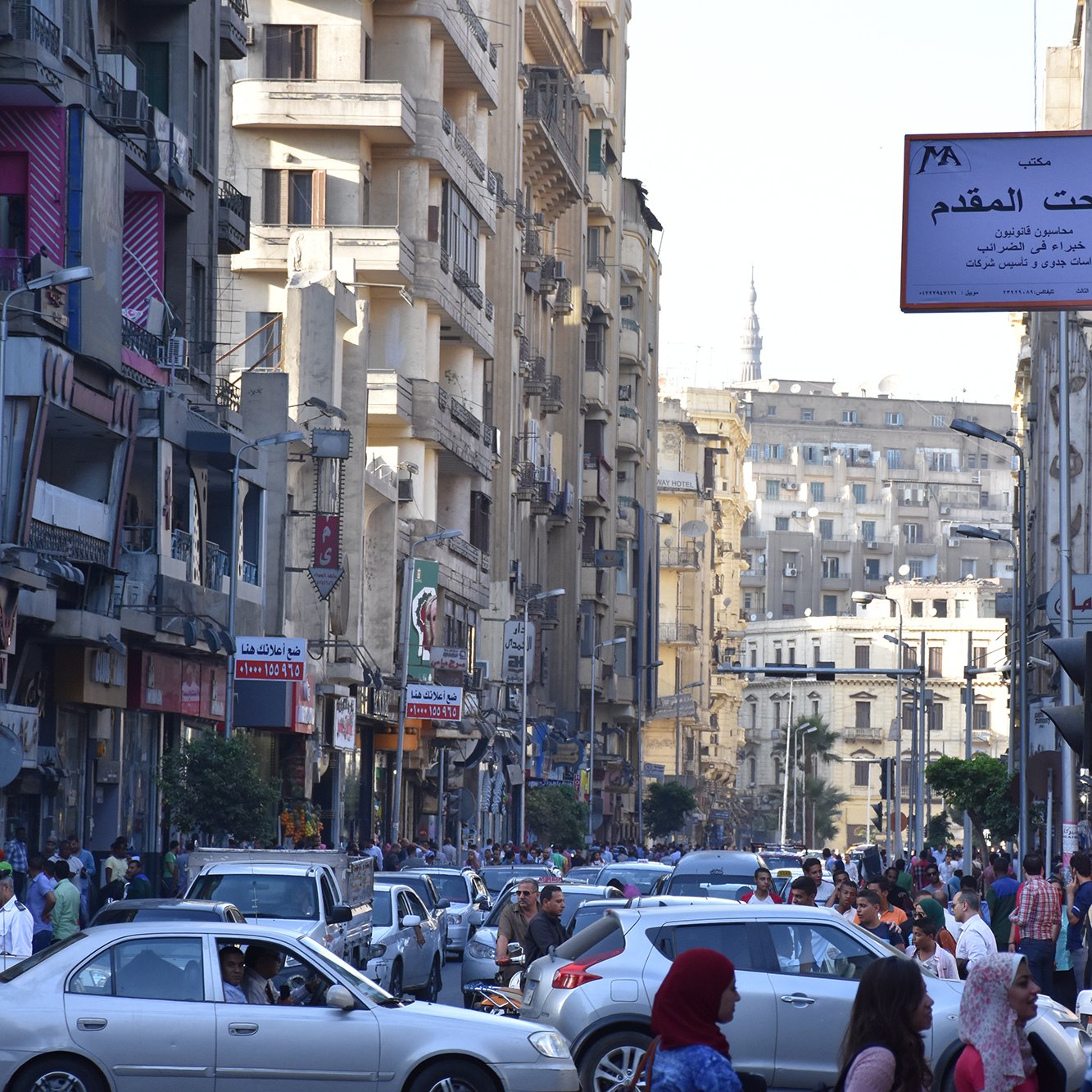 L'image montre une rue animée dans une ville, probablement une métropole. On peut voir de nombreux bâtiments de plusieurs étages qui bordent la route. Les voitures sont garées et circulent, créant un certain embouteillage. Une foule de personnes se déplace le long des trottoirs. Des panneaux de signalisation et des enseignes en arabe sont visibles, indiquant que cette scène se déroule dans un pays arabophone. L'atmosphère semble dynamique et vivante, typique d'une grande ville.