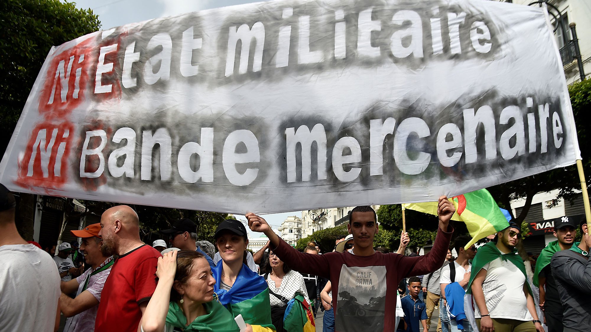 The image shows a large group of people participating in a protest or march. In the foreground, a man is holding a prominent banner that reads "Ni Etat Militaire Ni Bande Mercenaire," which translates to "Neither Military State Nor Mercenary Band." The crowd appears diverse, with individuals carrying flags and wearing various clothing styles, indicative of a collective demonstration. The background features urban scenery, suggesting the protest is taking place in a city.