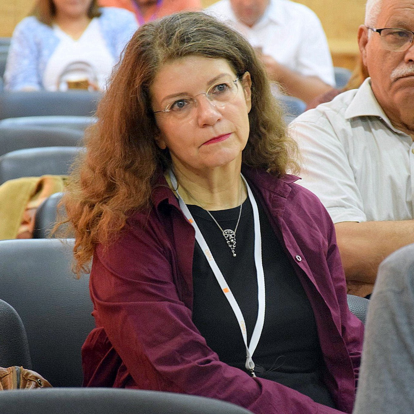 L'image montre une femme assise dans une salle de conférence ou de réunion, avec une expression pensive. Elle porte un haut de couleur sombre et a de longs cheveux bouclés. En arrière-plan, on peut voir d'autres personnes dans une atmosphère assez calme. La femme semble concentrée sur ce qui se passe autour d'elle.