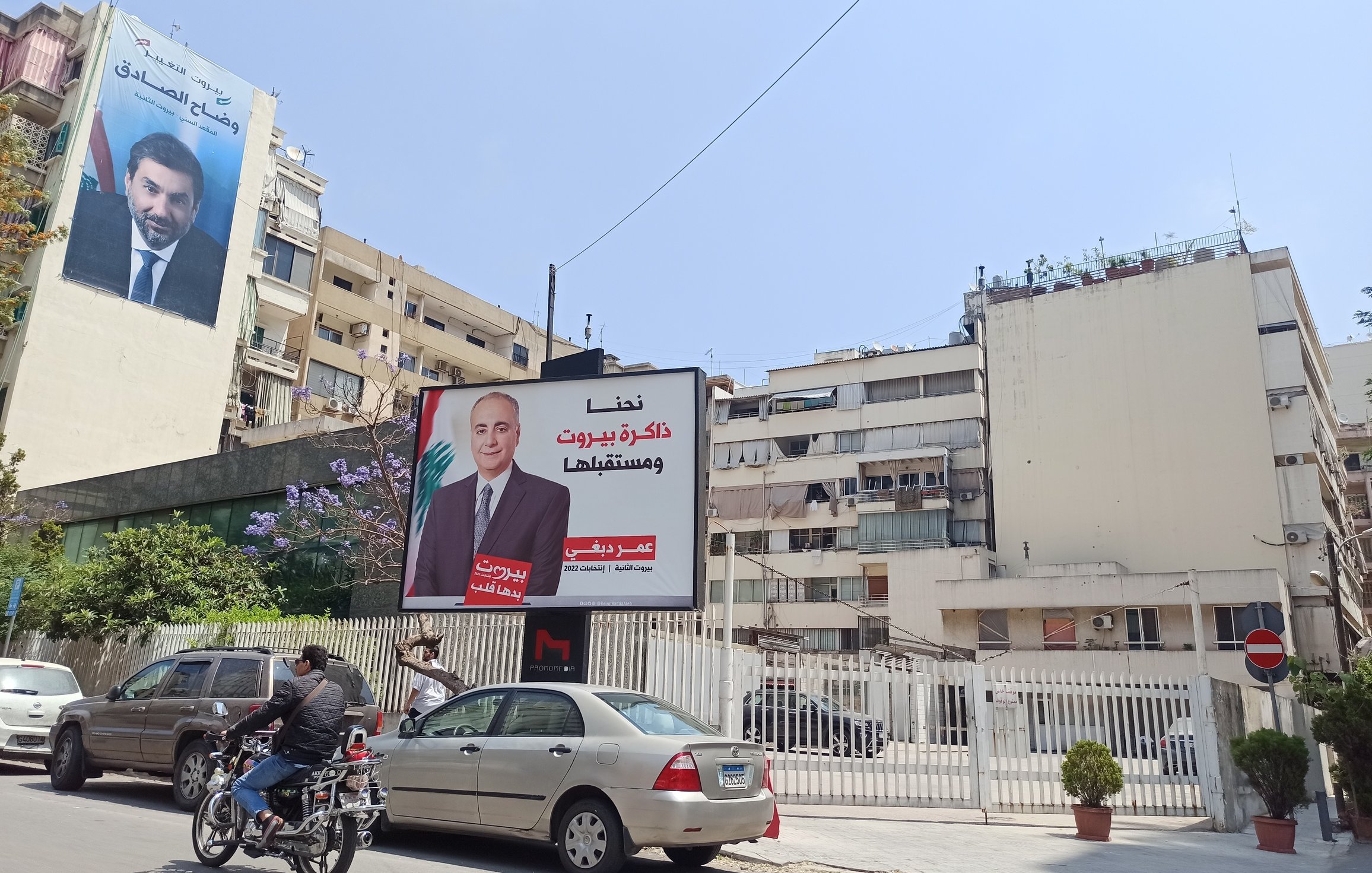 L'image montre une rue animée avec des voitures et des motos en circulation. À gauche, il y a une grande affiche sur un bâtiment, probablement une campagne politique, mettant en avant une personne, avec des textes en arabe. À côté, un autre bâtiment est visible, avec une façade claire et quelques plantes qui ajoutent une touche de verdure à l'environnement urbain. Le ciel est bleu, ce qui donne une ambiance ensoleillée à la scène.