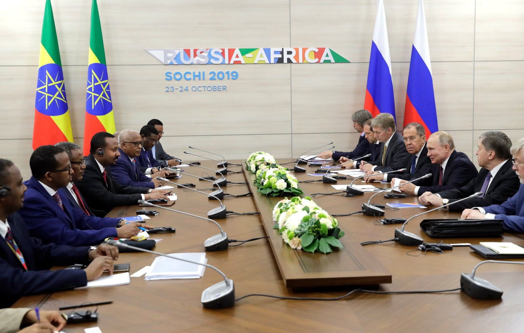 L'image montre une réunion officielle tenue à Sochi, en Russie, en octobre 2019, dans le cadre d'un sommet Russie-Afrique. On peut voir des représentants de plusieurs pays africains, ainsi que des dignitaires russes, assis autour d'une grande table de conférence. Des drapeaux représentant l'Éthiopie et un autre pays africain sont visibles en arrière-plan. Au centre de la table, il y a des arrangements floraux. Les participants semblent engagés dans une discussion, avec certains utilisant des écouteurs pour la traduction.