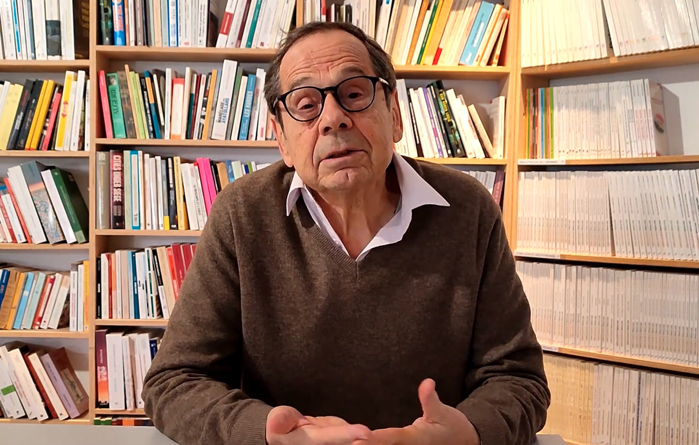 L'image montre un homme assis à une table, s'exprimant avec des mains gesticulantes. Il porte un pull brun et une chemise blanche. Derrière lui, une bibliothèque bien fournie avec des livres de différentes tailles et couleurs crée un environnement studieux et accueillant. L'homme semble être en train de partager des idées ou des réflexions.
