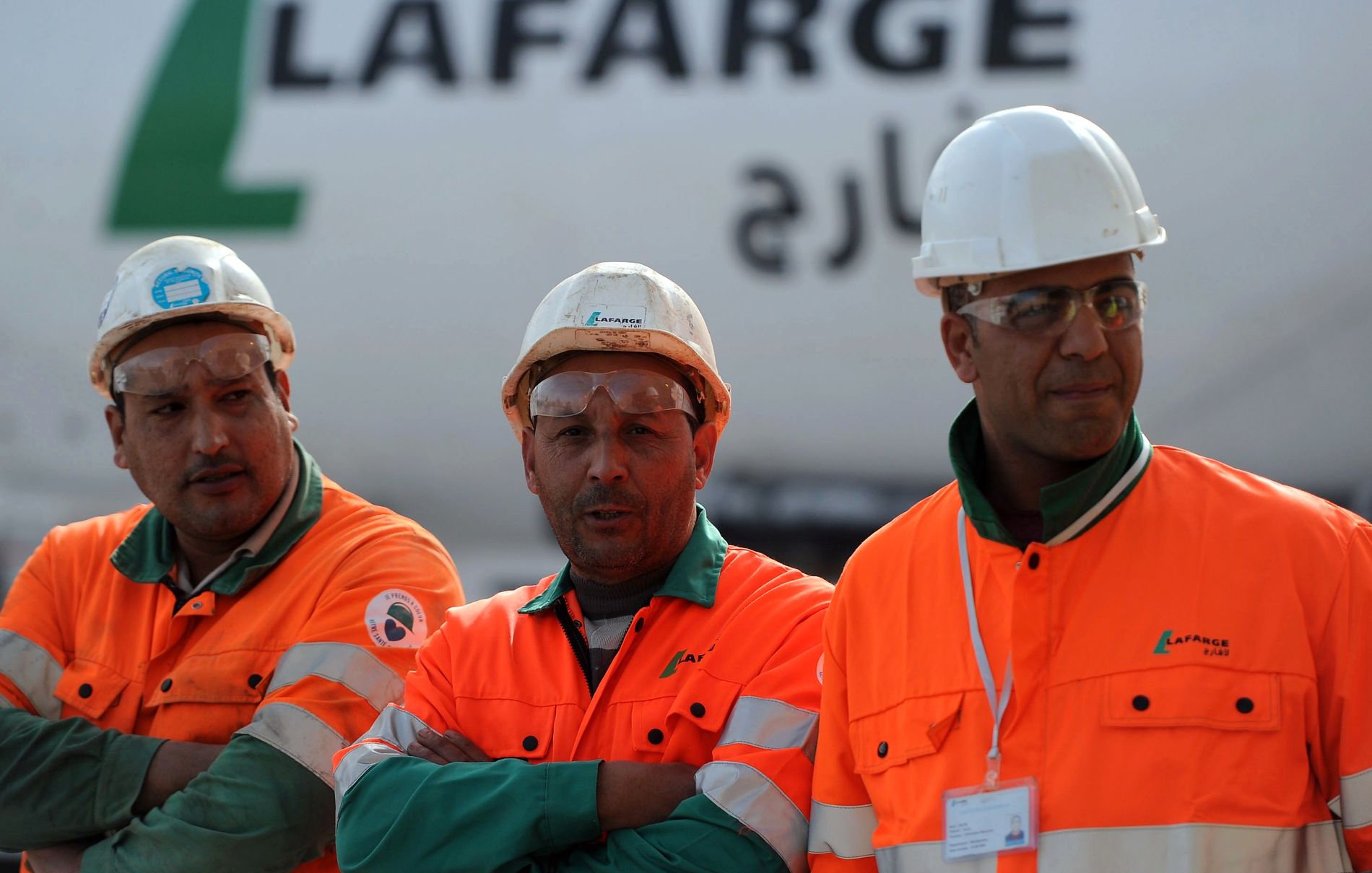 تظهر الصورة ثلاثة رجال يرتدون ملابس العمل البرتقالية، ويضعون خوذات بيضاء على رؤوسهم. يبدو أنهم يعملون في موقع بناء أو مصنع، حيث خلفهم تظهر شاحنة أو معدات تحمل اسم "لافارج". يعبر تعبيرهم عن الجدية والتركيز في العمل، بينما يقفون معًا.