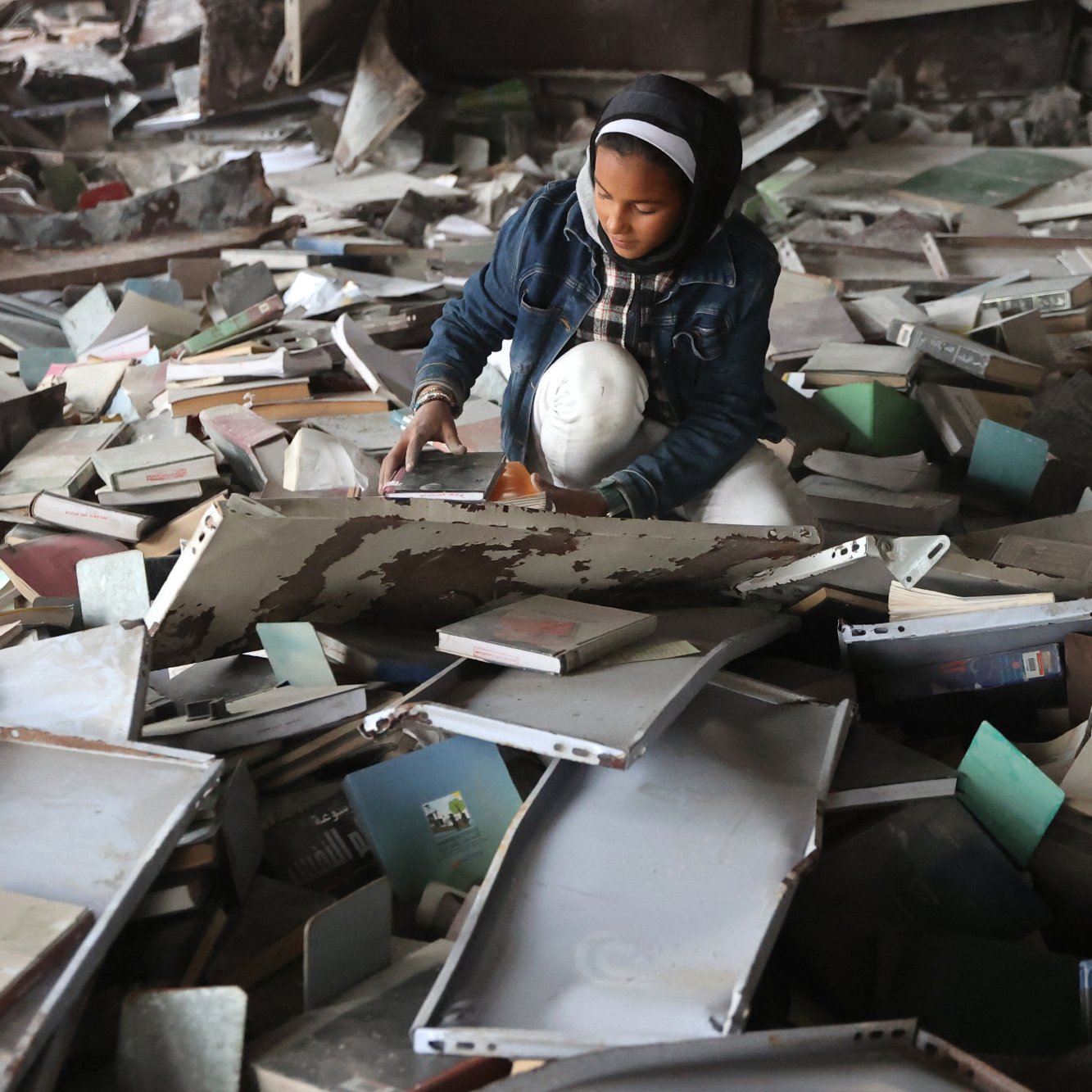 L'image montre une jeune fille assise au milieu d'un tas de livres et de débris. Elle semble explorer et manipuler les objets autour d'elle dans un environnement en désordre.