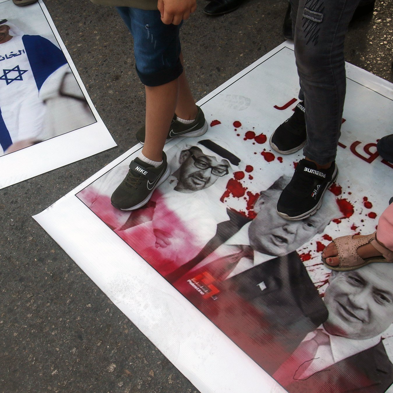 در این تصویر، تعدادی از افراد با پا بر روی پوسترهایی که عکس‌هایی از شخصیت‌های سیاسی معروف را نشان می‌دهند، ایستاده‌اند. یکی از پوسترها به تصویر نتانیاهو مربوط می‌شود و دیگری شامل تصاویری از ترامپ و یک شخصیت دیگر است. نشان‌های خون و نوشته‌ها بر روی پوسترها به وضوح دیده می‌شوند و این تصویر به نظر می‌رسد در یک تظاهرات یا اعتراض گرفته شده باشد.