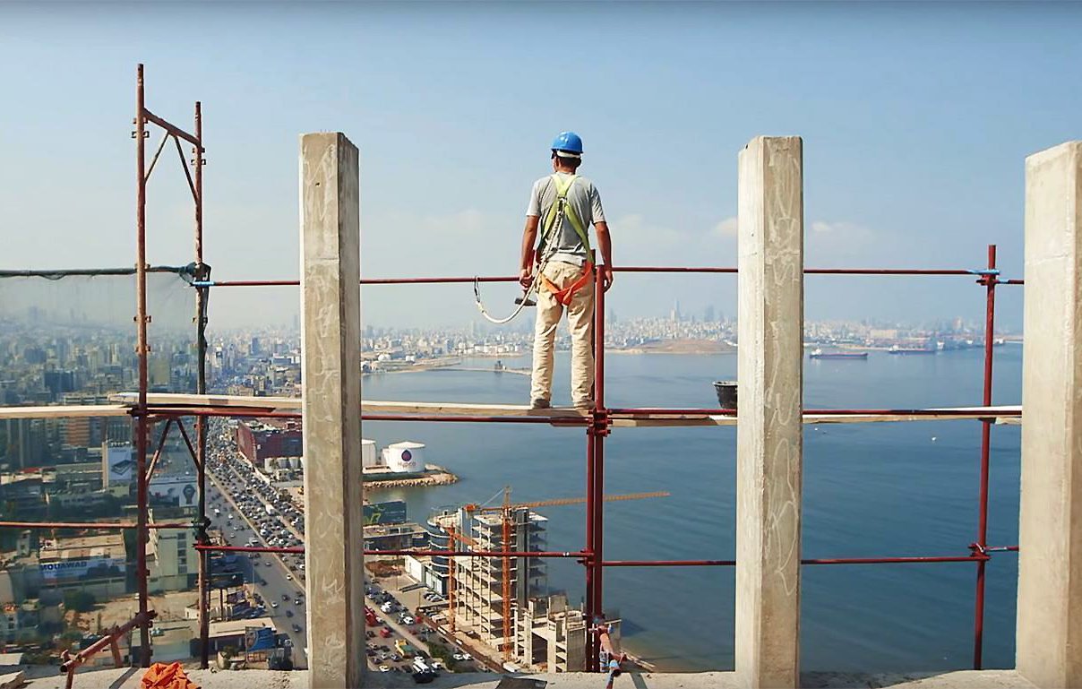 L'image montre un ouvrier de la construction se tenant sur un échafaudage, surplombant une vue panoramique sur une ville côtière. Il porte un casque de sécurité et des vêtements de travail. À l'arrière-plan, on peut voir des immeubles, une route qui longe la mer, ainsi que des bateaux sur l'eau. Le ciel est dégagé, laissant entrevoir une belle journée. La scène évoque à la fois le travail ardu des ouvriers et une magnifique perspective sur l'urbanisme et la nature environnante.
