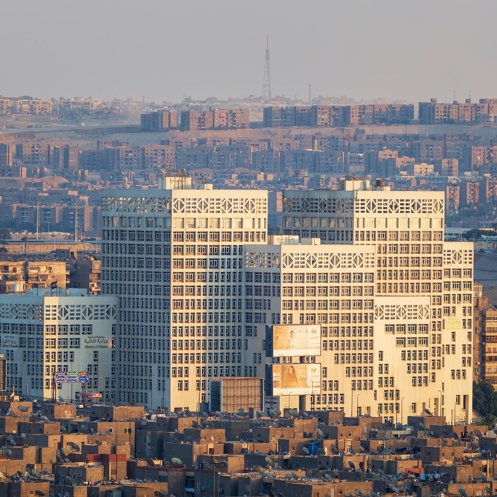 La imagen muestra una vista panorámica de una ciudad con edificios modernos en primer plano. Los edificios tienen un diseño arquitectónico distintivo y están iluminados por la luz del sol, lo que resalta su estructura. Al fondo, se pueden ver más edificios y una serie de montañas o colinas, lo que sugiere un paisaje urbano extenso. La atmósfera es clara, y la escena transmite una sensación de desarrollo urbano y modernidad.