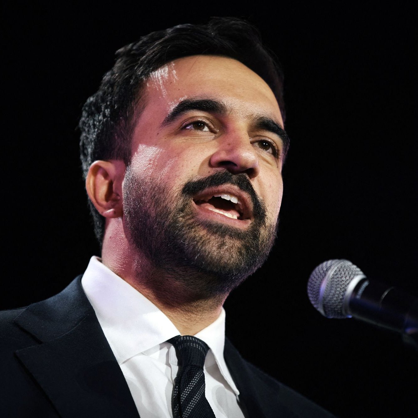 Homme en costume noir, parlant au micro avec une expression engagée.