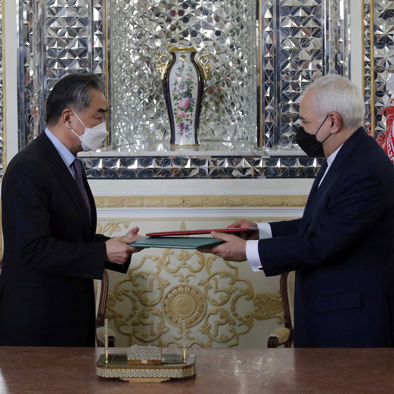 در این تصویر، دو دیپلمات در حال تبادل یک سند مهم هستند. در سمت چپ پرچم چین و در سمت راست پرچم ایران قرار دارد. هر دو فرد ماسک بر صورت دارند و به‌خاطر شیوع بیماری‌های تنفسی احتیاط می‌کنند. میز که بر روی آن سند قرار دارد، به‌نظر زیبا و مزین است. پس‌زمینه شامل تزئینات پیچیده و یک گلدان نیز هست که به فضای رسمی و دیپلماتیک این دیدار افزوده است.