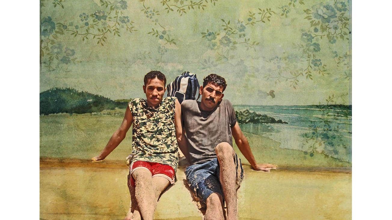 L'image représente deux jeunes hommes assis sur une plage. Ils portent des vêtements d'été ; l'un d'eux est en maillot de bain rouge et l'autre en shorts. Leur posture décontractée et le fond de plage évoquent une ambiance estivale. En arrière-plan, on peut voir des éléments naturels tels que l'eau et un paysage côtier, le tout stylisé par une texture artistique et des motifs floraux.