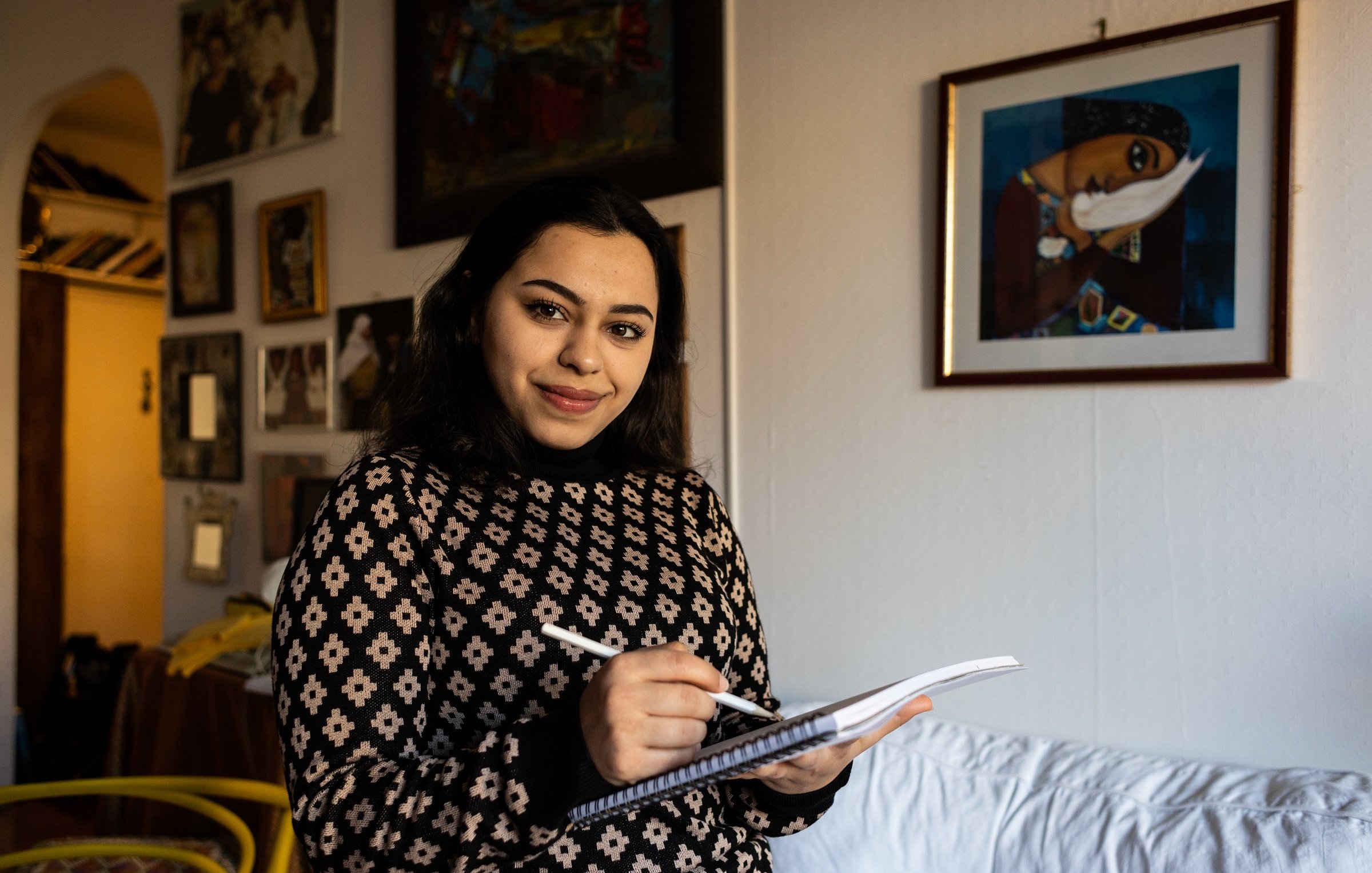 Nell'immagine, una giovane donna sorride mentre tiene un taccuino e una penna in mano. Indossa un maglione con un motivo geometrico. Sullo sfondo si può vedere una stanza con pareti decorate da vari quadri e fotografie. C'è un divano bianco e una sedia gialla, che aggiungono un tocco di colore all'ambiente. L'atmosfera è calda e accogliente.