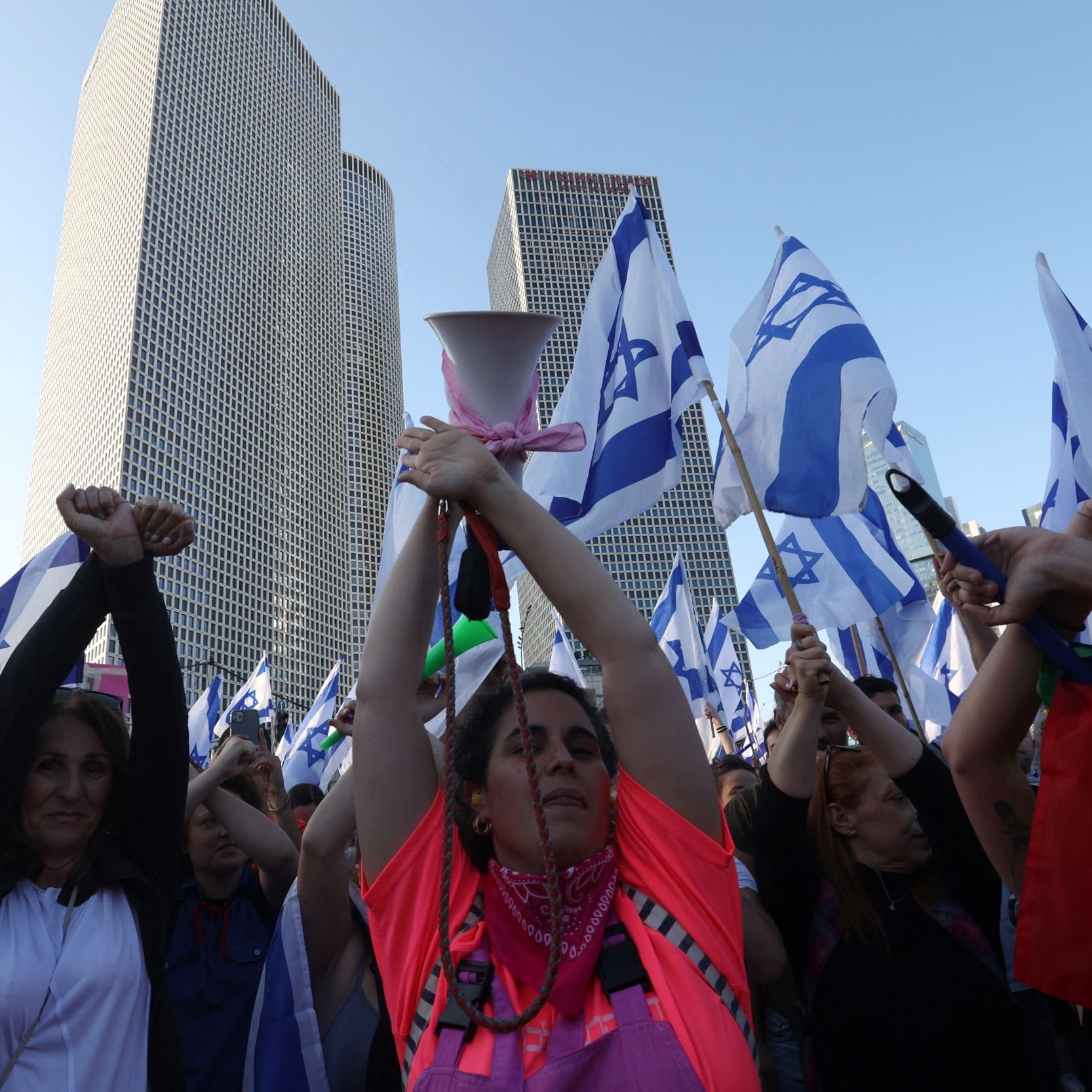 L'immagine mostra una folla di persone che partecipa a una manifestazione. Alcuni partecipanti tengono in alto delle bandiere israeliane, mentre molti altri alzano le mani, dimostrando un senso di unità e protesta. In primo piano, si può vedere una donna con una maglietta di colore neon che sembra guidare la manifestazione con un megafono. Sullo sfondo, si notano alti edifici moderni, suggerendo che l'evento si svolge in un contesto urbano. L'atmosfera è carica di energia e determinazione.