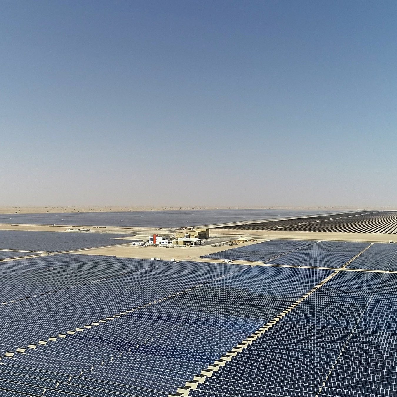 تظهر الصورة مزرعة شمسية كبيرة تحتوي على عدد كبير من الألواح الشمسية المنتشرة على مساحة واسعة. الألواح مرتبة في صفوف متوازية وتمتد إلى الأفق تحت سماء صافية وخالية من الغيوم. في الخلفية، يمكن رؤية محطة توليد الطاقة أو منشأة صغيرة تابعة للمزرعة، مما يدل على استغلال الطاقة الشمسية في توليد الكهرباء. تعكس الألواح الشمسية أشعة الشمس، مما يبرز نطاق المشروع وأهميته كجزء من استراتيجيات الطاقة المتجددة.