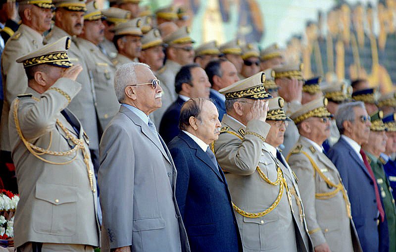 L'image montre un groupe de personnalités militaires en uniforme, saluant avec respect. Ils se tiennent debout en rangs, avec un décor cérémoniel en arrière-plan. Les hommes portent des uniformes ornés de médailles et de chapeaux militaires, et l'atmosphère semble officielle et solennelle, probablement lors d'une célébration ou d'un événement commémoratif. Des fleurs sont visibles au premier plan, ajoutant à l'ambiance formelle de la scène.
