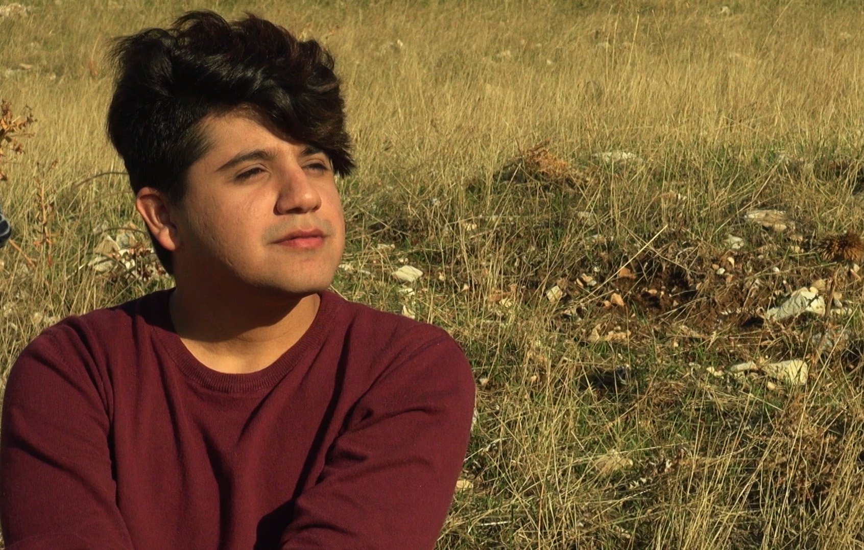 L'image montre une personne assise dans un champ de herbes hautes. La personne a les cheveux foncés et bouclés, et elle semble pensive, regardant au loin. Elle porte un pull de couleur bordeaux. L'environnement est naturel, avec une lumière douce, ce qui crée une atmosphère tranquille.