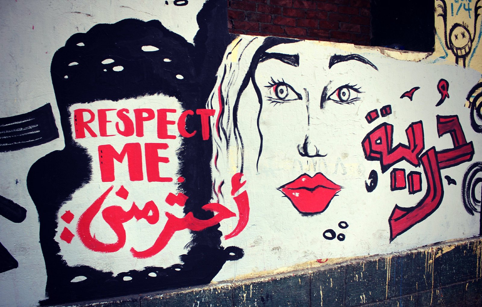 L'image montre une fresque murale qui combine des éléments graphiques et des mots en anglais et en arabe. À gauche, on peut voir le texte "RESPECT ME" en anglais, et en dessous, "احترمني" qui signifie "Respecte-moi" en arabe. À droite, il y a un visage stylisé de femme, avec des yeux expressifs et des lèvres rouges. Le tout est peint sur un mur de couleur claire, créant un message fort sur l'importance du respect.