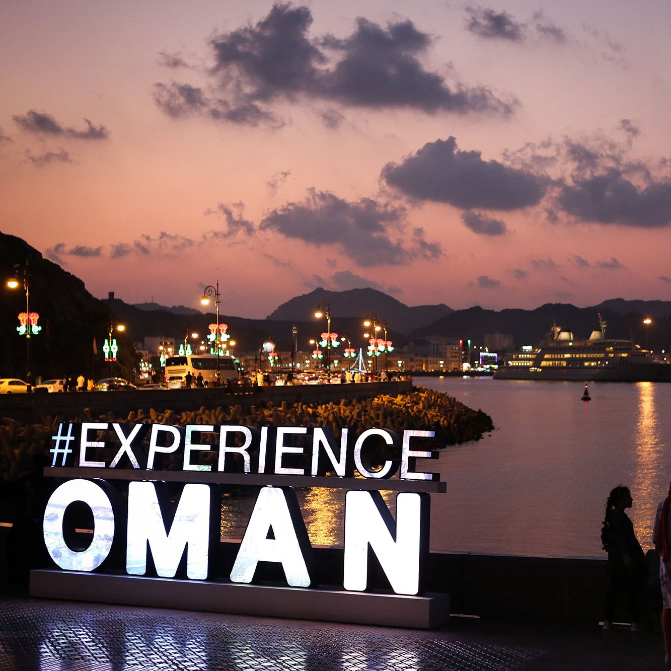 تظهر الصورة مشهدًا جذابًا يغرب فيه الشمس، حيث ينعكس الضياء الناعم على المياه. في المقدمة، يوجد شعار مضيء يحمل عبارة "#EXPLORE OMAN" مع خلفية جبلية وسماء ملونة تلقي بظلالها على البحر. يبدو أن هناك مجموعة من الأشخاص يتجمعون بالقرب من الشعار، في حين تظهر السفن في الميناء في الخلفية. الأجواء تبدو ممتعة ويدل المشهد على جمال عمان وطبيعتها الخلابة.