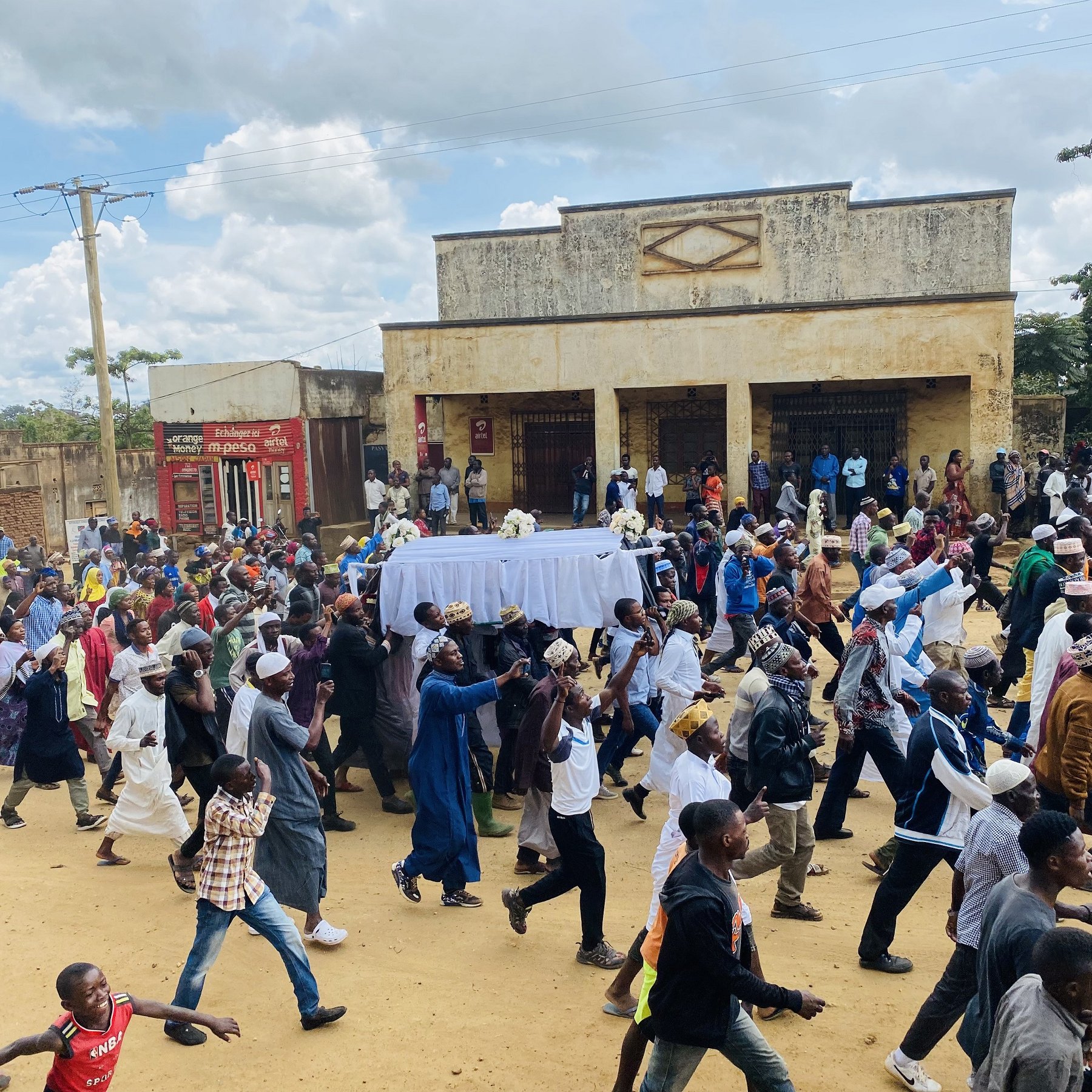 L'image montre une grande foule de personnes marchant dans une rue, probablement lors d'un événement ou d'une procession. Au centre, il y a un chariot couvert d'un drap blanc, et les gens semblent marcher avec une certaine ferveur. On peut apercevoir différents styles vestimentaires, suggérant une diversité parmi les participants. En arrière-plan, des bâtiments sont visibles, ainsi qu'un ciel partiellement nuageux. L'atmosphère semble collective et déterminée.