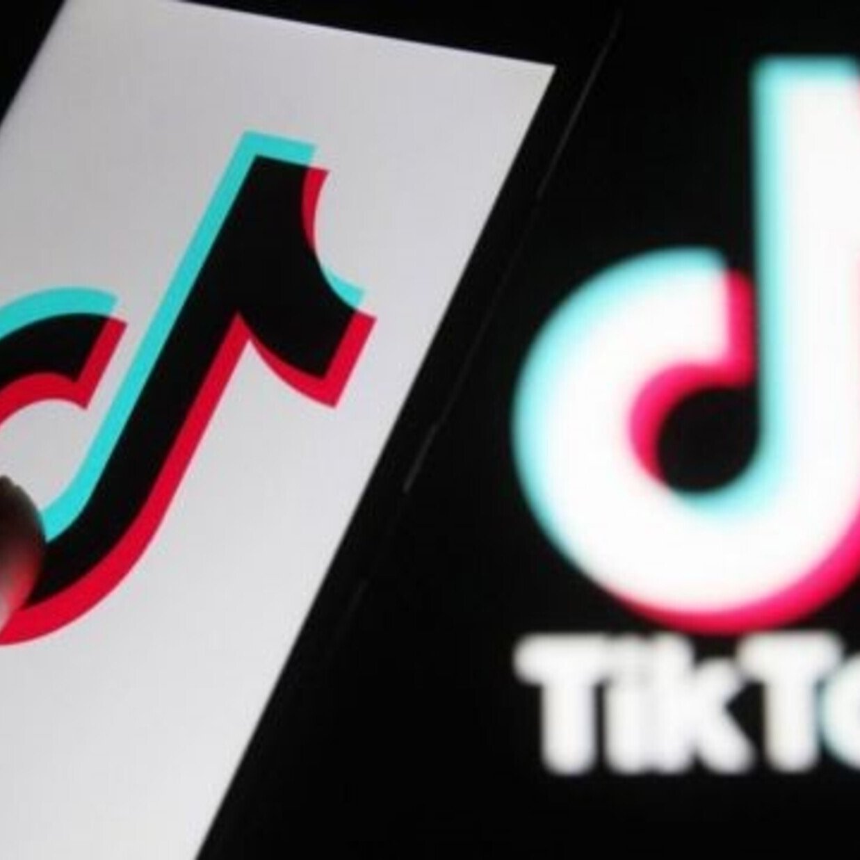 The image features a close-up of a smartphone displaying the TikTok logo, which is designed with vibrant colors—predominantly red, blue, and white. A finger is pointing towards the logo on the screen, while a larger version of the TikTok logo is blurred in the background. The overall focus is on the application's recognizable branding.