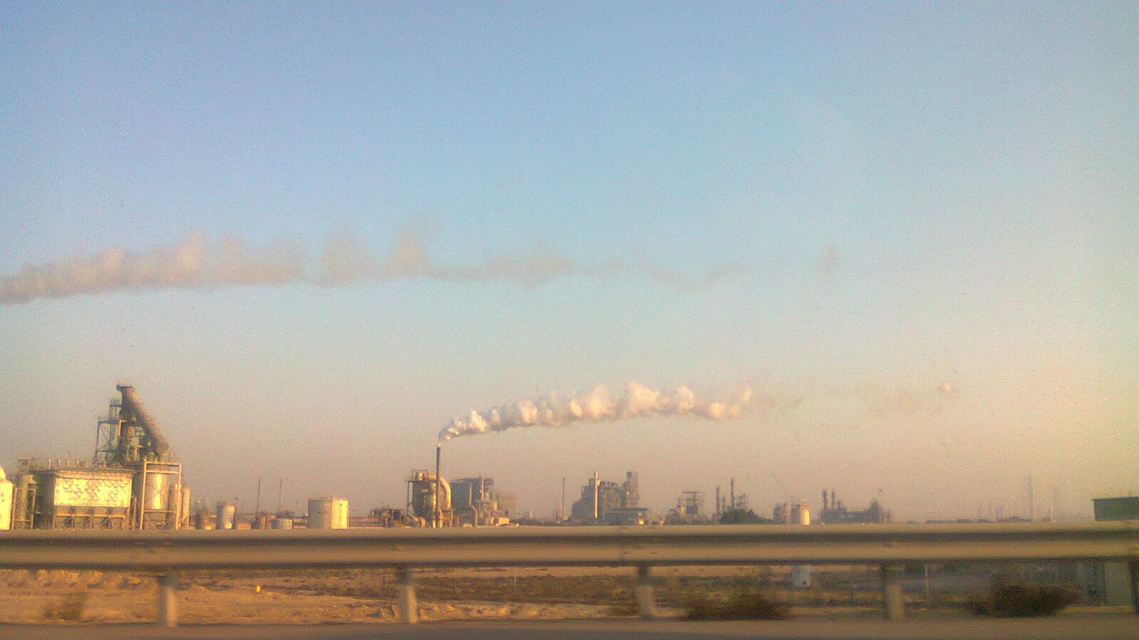 L'image montre un paysage industriel avec plusieurs structures d'usine. On voit de grandes cheminées qui émettent des nuages de fumée blanche dans le ciel. Le ciel est clair, avec une lumière douce, ce qui donne une atmosphère tranquille malgré l'activité industrielles. Au premier plan, on aperçoit une route avec une barrière en béton, indiquant que l'on observe ce paysage en passant en véhicule. L'ensemble dégage une impression de production industrielle dans un environnement ouvert.