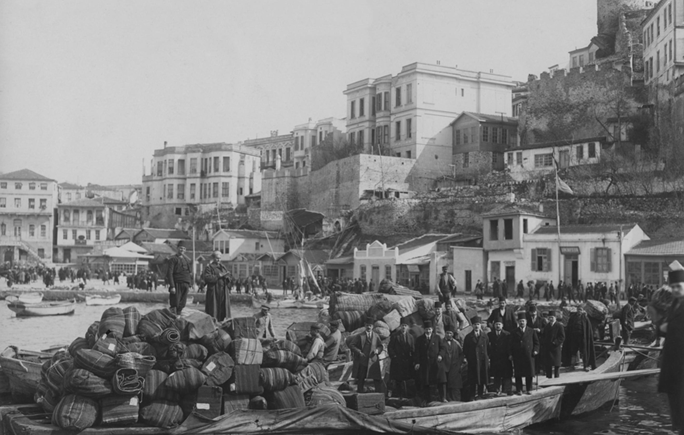 تظهر الصورة مشهداً تاريخياً من منطقة ساحلية، حيث توجد مجموعة من الأشخاص يقفون على قارب محمّل بأكياس وصناديق، مما يدل على حركة تجارية أو نقل للبضائع. في الخلفية، يمكن رؤية مبانٍ متعددة الطوابق وبعض الأسطح، بالإضافة إلى قلعة أو حصن يبدو أنه في حالة جيدة. هناك مجموعة من الناس تسير على الرصيف، مما يدل على النشاط والحركة في المنطقة. الأجواء العامة تعكس فترة زمنية سابقة، ربما أوائل القرن العشرين، مع تنوع في الملابس التي تشير إلى زمن بعيد.