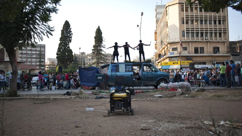 L'image montre une scène animée dans un endroit urbain. Au premier plan, on voit un espace dégagé avec un générateur. En arrière-plan, un certain nombre de personnes sont rassemblées, probablement pour assister à un événement. Sur le toit d'une fourgonnette bleue, trois personnes sont perchées, probablement en train de performer ou de s'exprimer de manière artistique. L'environnement semble être un mélange de vie urbaine et de rassemblement communautaire.