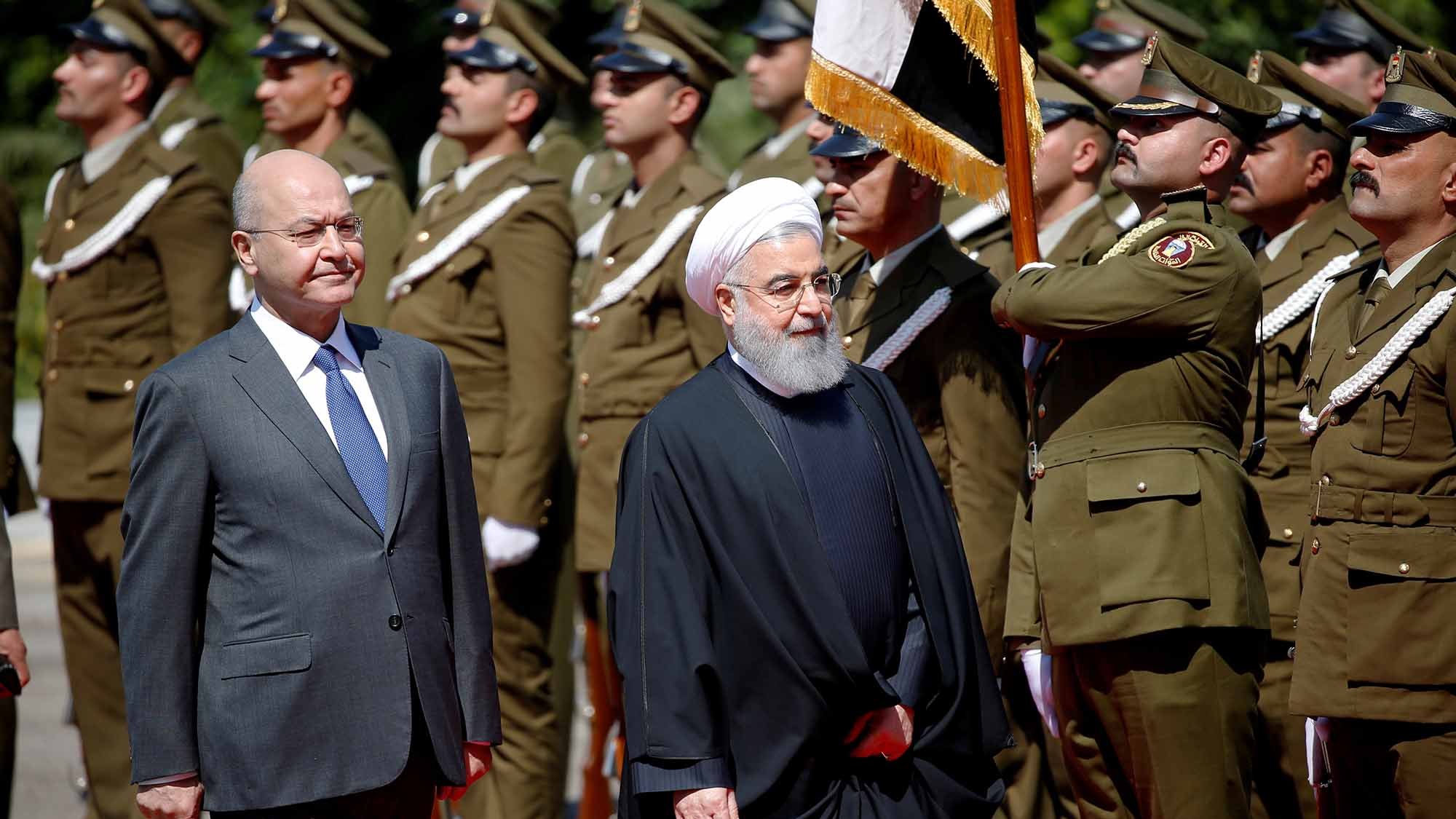 در این تصویر، دو شخصیت در حال برگزاری یک مراسم رسمی هستند. در سمت چپ، فردی با لباس رسمی و کت و شلوار ایستاده است. در سمت راست، فرد دیگری با لباس سنتی ایرانی، که به یک عمامه و عبای سیاه پوشیده است، حضور دارد. در پس‌زمینه، تعدادی از نظامیان لباس‌پوشیده به خط ایستاده‌اند و در حال انجام تشریفات نظامی هستند. تصویر نشان‌دهنده یک رویداد رسمی یا دیدار دیپلماتیک است.