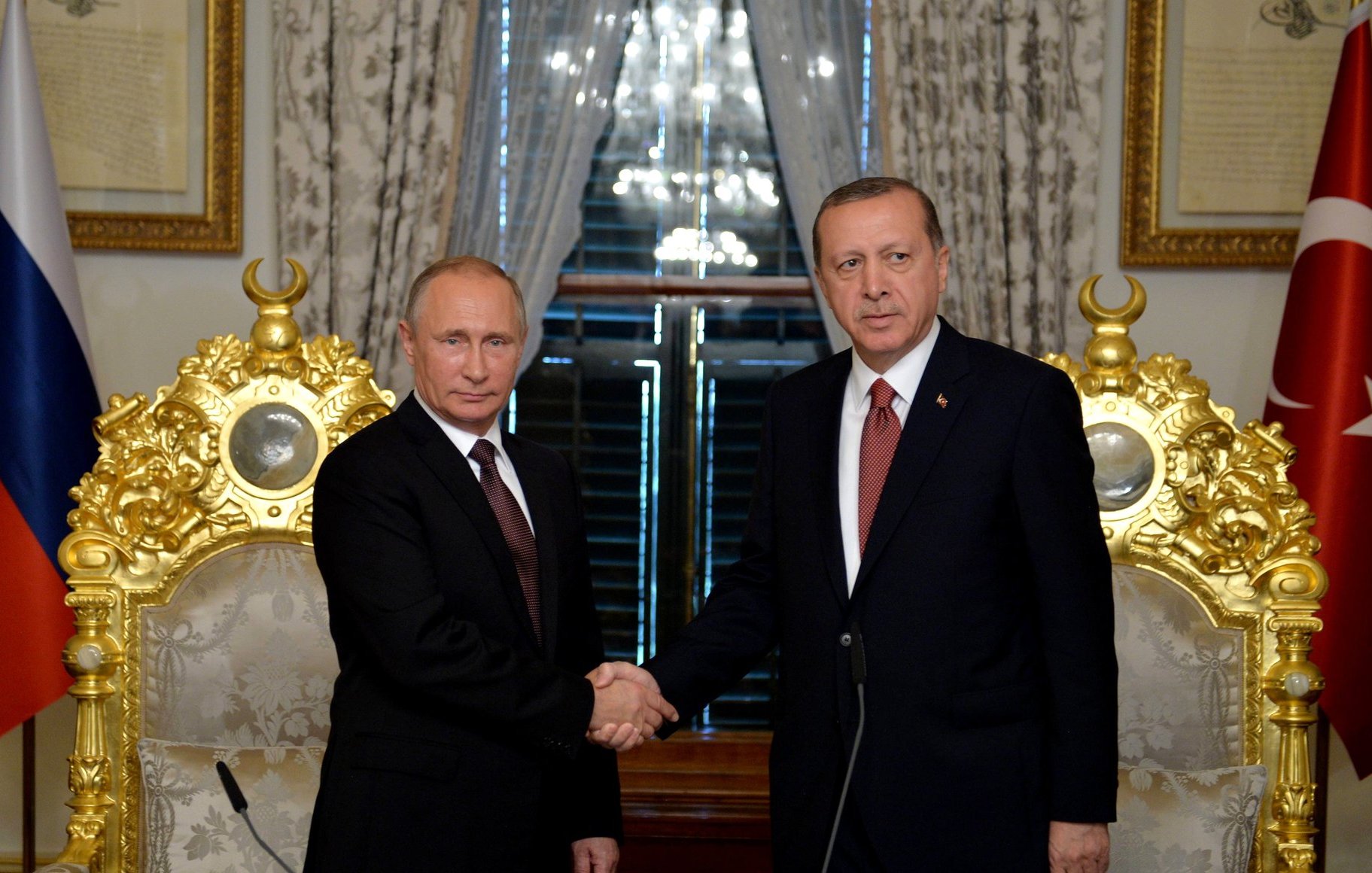 L'image montre deux hommes se serrant la main dans un cadre formel. Ils sont probablement en train de discuter d'accords ou de collaborations. L'un d'eux semble être en costume sombre avec une cravate, tandis que l'autre porte également un costume et une cravate. Ils se tiennent devant des fauteuils élégants et ornés, avec des drapeaux en arrière-plan, ce qui suggère un contexte officiel ou diplomatique. La lumière naturelle entre par les fenêtres, créant une atmosphère importante.