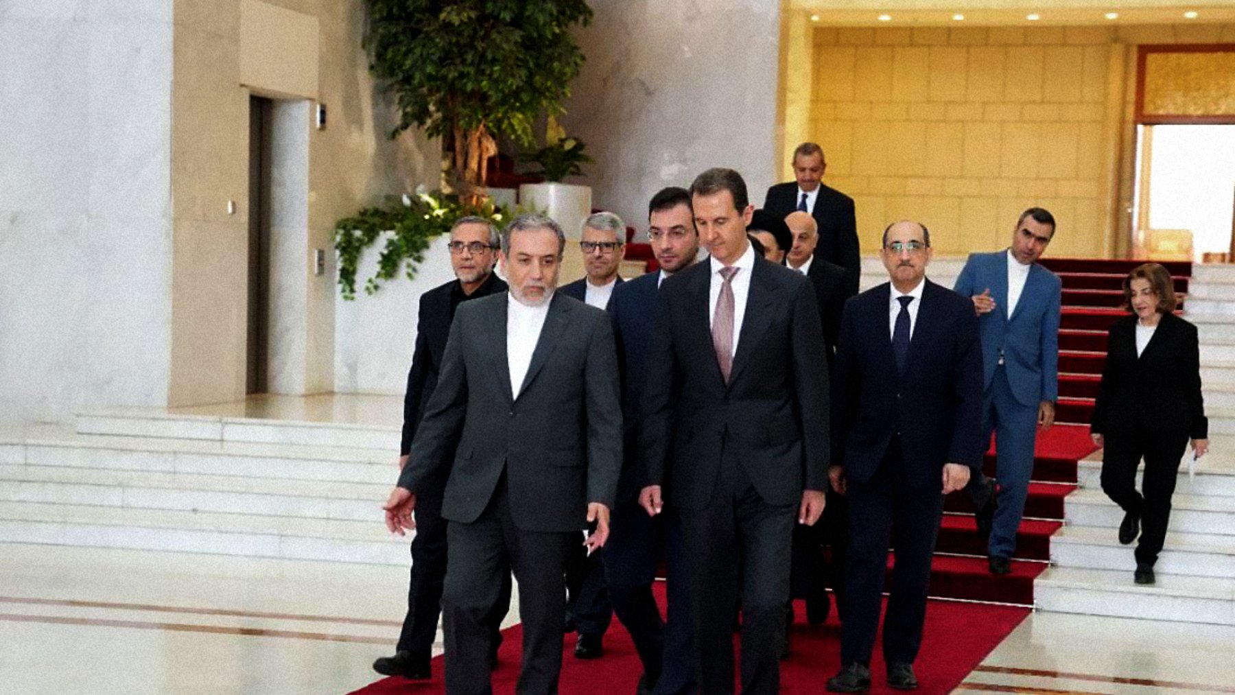 L'image montre un groupe de personnes marchant sur un tapis rouge dans un hall d'entrée élégant. À l'avant, deux hommes semblent en conversation, tandis que d'autres personnes les suivent, allant d'une manière formelle. L'environnement est spacieux avec des murs clairs et une décoration soignée, indiquant un lieu officiel ou diplomatique.