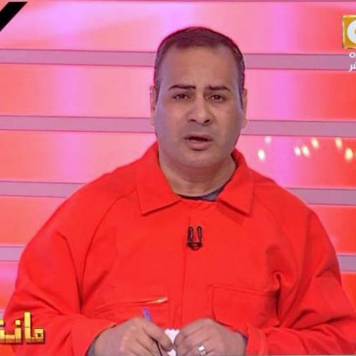 L'image montre un homme en train de parler devant un fond rouge avec des lignes lumineuses. Il porte une combinaison orange et semble être en train de faire une présentation ou de commenter quelque chose. Son expression faciale est sérieuse et il tient un stylo dans sa main. Le décor et son apparence suggèrent qu'il s'agit d'un animateur ou d'un commentateur à la télévision.