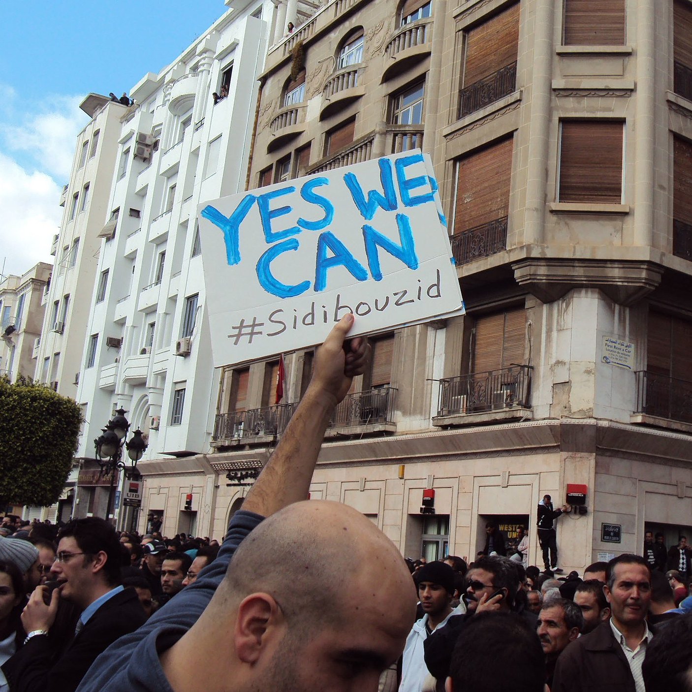 در این تصویر، گروهی از مردم در یک تجمع یا تظاهرات شرکت کرده‌اند. شخصی در جلوی تصویر، تابلویی با نوشته «YES WE CAN» و هشتگ «#Sidibouzid» را در دست دارد. ساختمان‌هایی با معماری مشخص در پس‌زمینه دیده می‌شود و جمعیت به‌نظر معترض یا فعال است. فضای تصویر حالت اجتماعی و سیاسی به خود گرفته و نشان‌دهندهٔ یک حرکت جمعی است.
