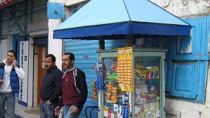 L'image montre un kiosque de vente situé sur un trottoir. Le kiosque, de style ancien, est peint en bleu et abrite divers articles comme des snacks, des boissons et des produits de consommation courante, visibles à travers les vitres. À côté du kiosque, il y a deux hommes. L'un porte un t-shirt, tandis que l'autre est habillé en noir. En arrière-plan, on peut voir des bâtiments aux murs usés et des volets bleus. L'atmosphère a l'air animée et typique d'un marché ou d'une rue commerçante.