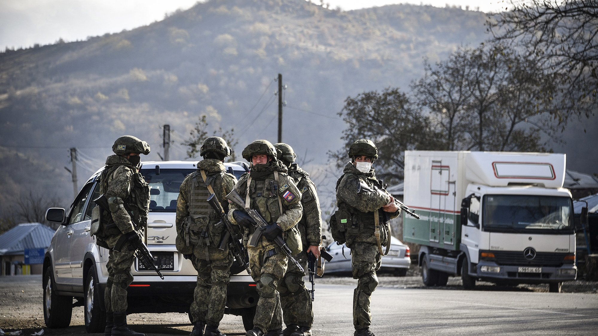 L'image montre un groupe de soldats armés en uniforme, se tenant près d'un véhicule. Ils semblent être en position de vigilance, avec des montagnes en arrière-plan. L'atmosphère est sérieuse, suggérant une opération militaire ou une situation de sécurité. Des camions peuvent également être aperçus à proximité, ajoutant un contexte logistique à la scène.