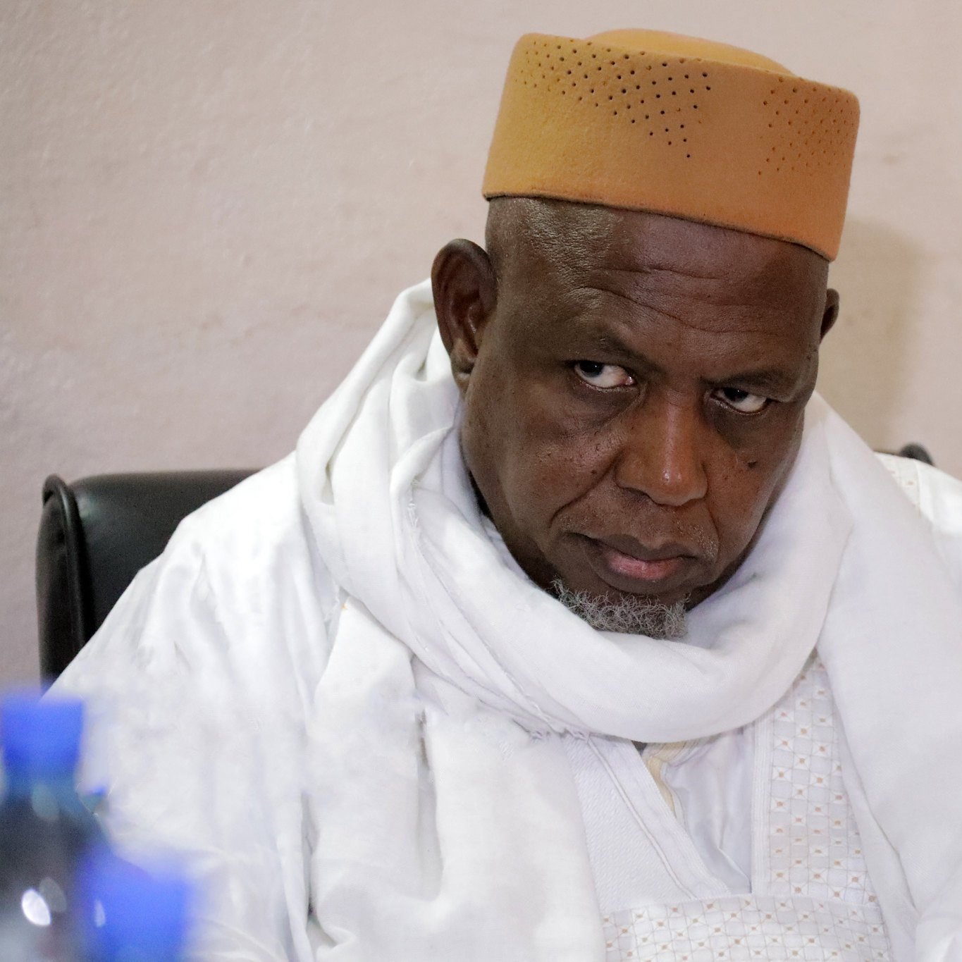 این تصویر یک مرد را نشان می‌دهد که لباسی سنتی به تن دارد و یک سرپوش قهوه‌ای رنگ بر سر گذاشته است. او در حال نشسته و به جلو خیره شده است، به نظر می‌رسد که در حال تفکر یا گوش دادن به صحبت‌های دیگران است. پس‌زمینه تصویری ساده و بدون هیچ جزئیات خاصی دارد.